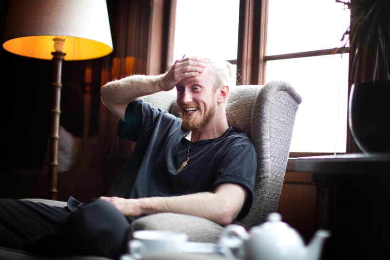 Fredrik Brännlund laughing with a hand to his forehead in an armchair by a window