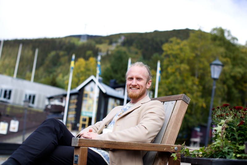 Fredrik Brännlund sitting on a wooden bench outdoors in a beige coat, a forested mountainside behind him